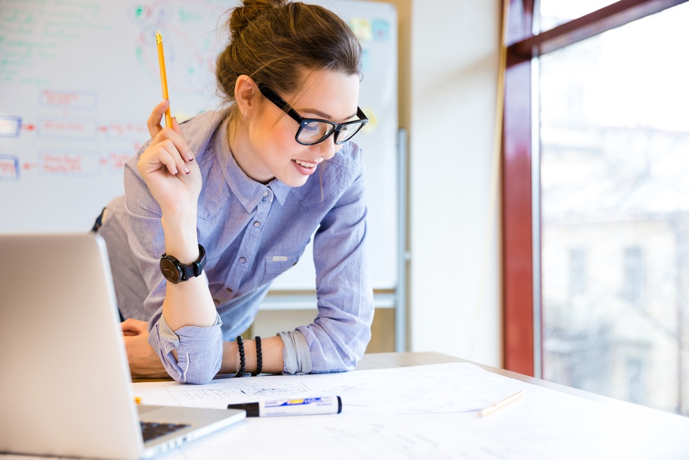 Happy young woman in glasses standing near the window in office and working with blueprint 